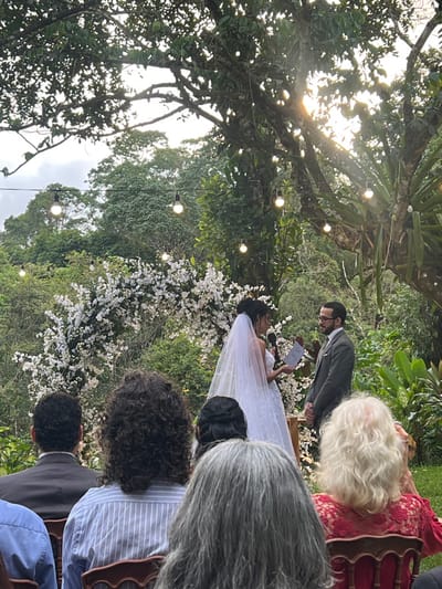 An outdoor wedding ceremony is depicted in Camaragibe, Brazil. The scene features a bride and groom standing at the center. The bride, wearing a white wedding gown and veil, holds a microphone in her left hand and papers in her right, appearing to speak. She faces the groom, who is dressed in a gray suit with a white shirt and dark tie, standing with his hands clasped in front of him. Behind the couple is a decorative arch adorned with white flowers. String lights with illuminated bulbs are strung through the surrounding trees, and sunlight filters through the dense foliage, suggesting late afternoon. The background consists of abundant green trees and lush vegetation, indicating a natural, forested, or garden environment. In the foreground, partially visible from behind, are several wedding guests seated on dark wooden chairs, observing the ceremony. Their backs and heads are visible, with varying hair colors and styles.