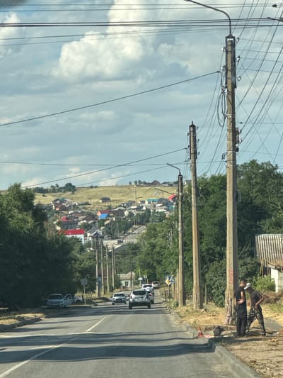 The image depicts a roadside scene in a rural area, likely in Russia given the location context. The orientation of the image is rotated, showing a street on the right side and a field with houses on the left, separated by a line of utility poles and trees. The sky is partly cloudy, suggesting daytime with fair weather.
In the foreground on the right side of the road, two individuals are visible. One person is standing, leaning against a structure, and another is seated. They appear to be engaged in some casual activity or waiting. Further down the road, several cars are driving or parked. One vehicle, a white SUV, is prominent. On the left side of the road, a field is dotted with houses, many with colorful roofs, indicating a residential area within a village or small town. The utility poles are a prominent feature, carrying numerous wires that crisscross the frame, suggesting a well-connected area despite its rural setting.
Notable details include the presence of traffic cones and construction barriers near the road, hinting at ongoing work or road maintenance. The overall impression is one of a normal day in a rural Russian settlement, with daily activities and infrastructure visible. No specific text is clearly legible in the image.