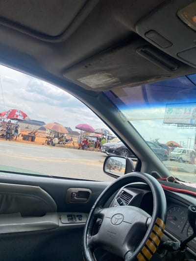 The image depicts a roadside scene in Naze, Nigeria, viewed from the interior of a vehicle, specifically from the driver's perspective. The foreground shows the vehicle's gray dashboard, a black steering wheel with a Toyota emblem and yellow-striped cover, the driver's side door panel with power window controls, and the rearview mirror reflecting a white van and a motorcycle.
Through the windshield, a busy street scene unfolds under an overcast sky. Multiple informal market stalls are visible along the road, characterized by large, colorful umbrellas providing shade, including red and white striped, brown, pink, and multi-colored patterns. Numerous individuals, appearing to be vendors and customers, are present near these stalls, engaged in various activities. Objects like wooden tables, benches, and merchandise are discernible at the stalls.
On the paved road, a dark-colored Hyundai sedan is parked or stationary in the midground, with other vehicles, including a white van, further down the road. The immediate roadside features patches of exposed dirt. In the background, behind the market activity, several low-rise buildings constructed with what appears to be reddish-brown bricks are visible.