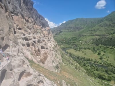 The image captures a dramatic cliffside dwelling, likely the Vardzia cave monastery complex in Aranisi, Georgia. The sheer rock face is carved with numerous caves and openings, resembling a honeycomb structure. Several levels of these ancient dwellings are visible, with pathways and stairs carved into the rock, some of which are lined with metal railings for safety. A few individuals, appearing to be tourists, are seen on these pathways and stairs, suggesting they are exploring the historical site.
The cliff overlooks a lush green valley, through which a dark river winds its way. The valley is dotted with trees and appears to be a rural area, with a few small buildings or houses visible in the distance. The surrounding landscape is characterized by rolling green hills and mountains, with a clear blue sky overhead, punctuated by a few scattered white clouds. The overall scene conveys a sense of history, nature, and human endeavor.