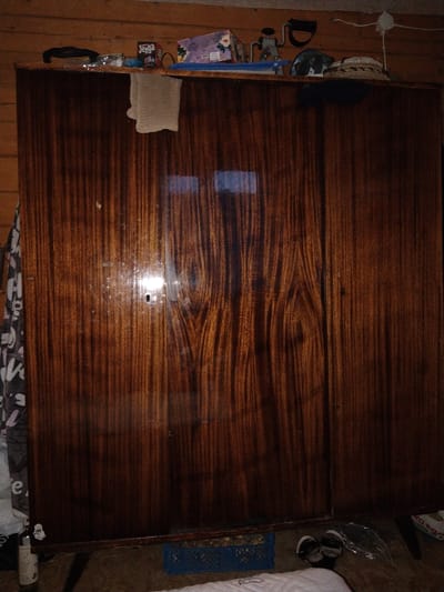 The image shows a close-up of a large, dark wooden wardrobe, likely located in a home in Brish, Russia. The wardrobe has a highly polished, glossy finish with prominent wood grain patterns, showcasing deep reds and browns. It appears to be a two-door unit, with a visible seam down the middle.
On top of the wardrobe, a collection of various objects are scattered. These include a box of Jenga, a small blue object, what looks like a bottle of cleaning spray, a black looped item, a gray cloth, a small metal clamp or tool, and a woven hat. The lighting suggests it might be daytime, but the scene is indoors, and the wood grain reflects the light, creating highlights and shadows. The overall impression is of a domestic, possibly cluttered, space. There is no explicit activity or event taking place. The visible text is limited to the "Jenga" logo on the game box.