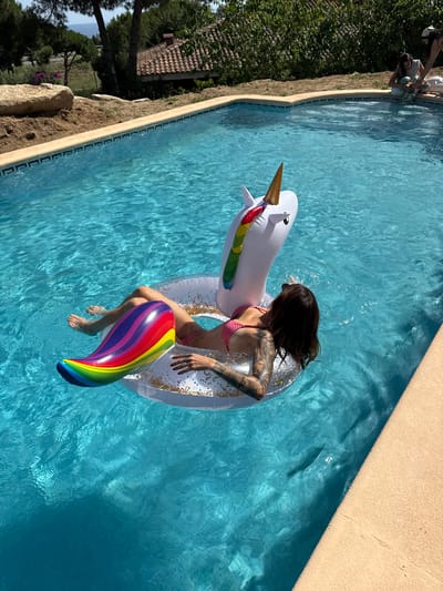 A woman with tattoos on her arm is floating on a unicorn-shaped inner tube in a swimming pool. She is wearing a red and white polka dot bikini and has a rainbow-colored fin emerging from her waist. The swimming pool is surrounded by a sandy-colored deck. In the background, there is a tiled roof house with trees and vegetation. The location is Sant Vicenç de Montalt, Spain.