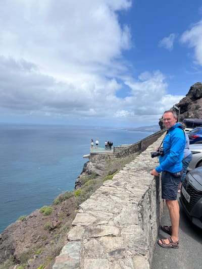 A medium shot depicts a man wearing a blue jacket and sandals leaning over a stone wall. In front of him is a camera. Behind him are parked cars. In the background, several people are standing on a glass platform. The image overlooks the ocean. The location is La Aldea de San Nicolás, Spain.