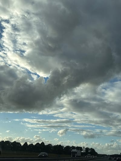 The image captures a highway scene near Staphorst, Netherlands, under a dramatic sky filled with both dark and light clouds. The perspective is from a vehicle in motion, looking forward. In the foreground, the highway is visible with several vehicles, including a white semi-trailer truck and a black car. A dark green sign is partially visible on the right side of the road. Beyond the highway, a line of trees forms a dark silhouette against the sky, with a few taller trees interspersed. The sky is the dominant feature, with large, textured clouds casting shadows and allowing patches of blue sky to show through. The overall impression is of a dynamic and somewhat overcast day, with the infrastructure of the highway contrasting with the natural beauty of the sky and the surrounding landscape.