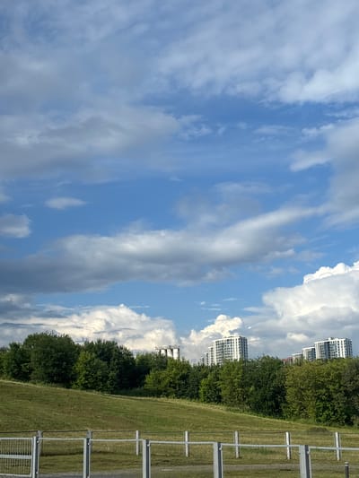 The image depicts a partially green, grassy hill in the foreground with a silver metal fence running horizontally across it. Behind the hill, a dense line of green trees forms a natural barrier. Beyond the trees, several modern, white, multi-story buildings are visible, indicative of a suburban or urban fringe area. The sky above is a vibrant blue, populated with a mixture of fluffy white cumulus clouds and darker, more scattered clouds, suggesting a dynamic weather system. In the far distance, several tall, cylindrical industrial structures can be seen. The overall scene suggests an outdoor environment with a blend of natural landscape and modern architecture, likely located in or near the Сеніцкі сельскі Савет region of Belarus. There are no people or animals visible in the image.