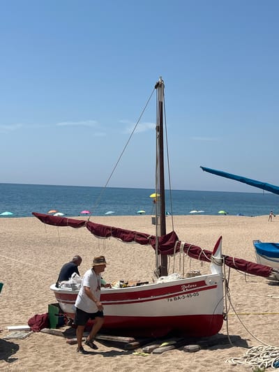 This media file shows a sailboat on a sandy beach. The sailboat is white with red accents and has the name "Dolors" and "7a BA-3-45-05" visible on its side. Two people are near the sailboat; one is wearing a white shirt and a hat, and the other is wearing a dark blue shirt. The scene appears to be a sunny day with a clear blue sky and calm blue water in the background. There are several colorful beach umbrellas scattered along the shore. The overall mood seems relaxed, possibly a vacation or leisure activity. The location is identified as Sant Pol de Mar, Spain.