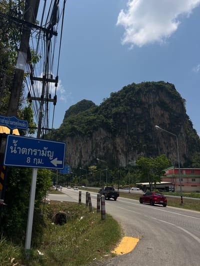 This is an outdoor photograph of a road scene in Tambon Takdat, Thailand. In the background is a large, rocky mountain covered with green vegetation. A two-lane road runs alongside the mountain, with a few vehicles including a red car and a dark-colored SUV on the road. Utility poles with tangled wires are visible on the left side of the frame.
A blue road sign in the foreground has white text in Thai script. Some of the text on the sign says "น้ำตกรามัญ 8 กม.", which translates to "Raman Waterfall 8 km". The sign also has a left-pointing arrow. Additional sign details are shown in the bottom left corner.
The weather appears to be sunny with a blue sky and scattered white clouds. The roadside is bordered by a grassy area with litter and a few small concrete barriers.