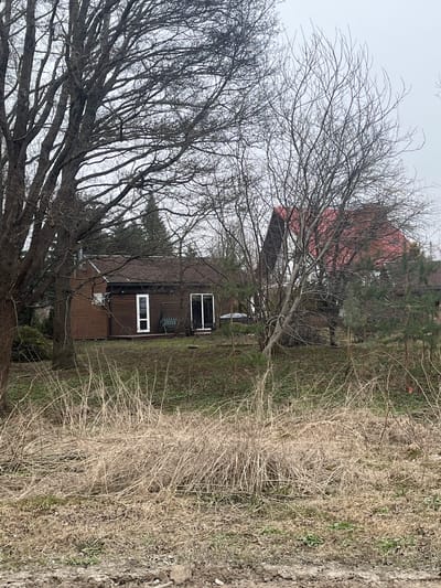 The image shows a rural scene in what appears to be Grīnvalti, Latvia. A brown, two-story house with white-framed windows is visible through bare trees. The roof is red. The foreground is dominated by dry, brown vegetation. The sky is overcast and grey. No people are visible.