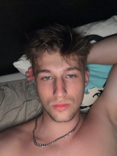 The image shows a close-up, overhead selfie of a young man lying down, seemingly in bed. He has light brown, messy hair and blue eyes. He is wearing a silver chain necklace. The background includes a pillow with a cartoonish face printed on it, a beige duvet, and a dark background. There is no visible indication of the city of Vanino, Russia in the image.