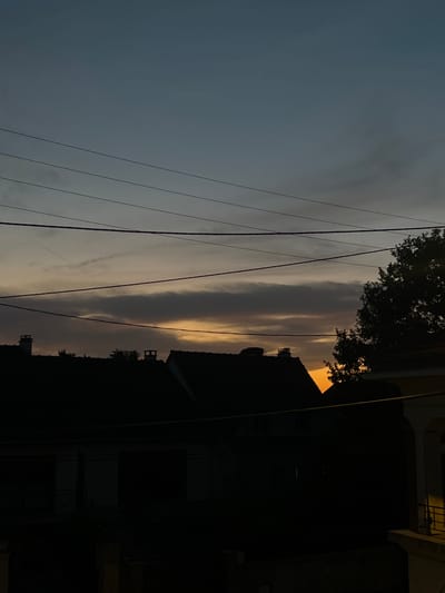 The image depicts a twilight scene over a residential area, likely Montfermeil, France. The sky is a gradient of deep blue to pale orange, with distinct cloud formations stretching horizontally across the frame. Several dark, silhouetted power lines are strung across the upper portion of the image. In the foreground, the dark silhouettes of rooftops are visible, some with chimneys. The harsh lighting of what appears to be a streetlamp illuminates the right side of the image, casting a yellow glow on a building's facade and a balcony railing. The lower portion of the image is very dark, obscuring details of the ground level and any potential inhabitants. The overall atmosphere is one of dusk settling over a quiet neighborhood.