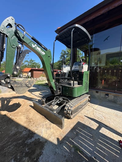An exterior, low-angle shot captures a green mini-excavator parked on a paved surface in front of a building with a large glass facade. The excavator is positioned slightly to the left of the frame, with its tracks and blade visible in the foreground. The boom and bucket of the excavator are extended, casting a long shadow on the ground. The building behind the excavator features a wooden-trimmed roof and a dark-tinted glass wall. A silhouette of a bird in flight is visible on the glass. The environment suggests a sunny day with a clear blue sky. The excavator is branded with "MÜLLER MACHINERY". The scene is located in Ribaritsa, Bulgaria.