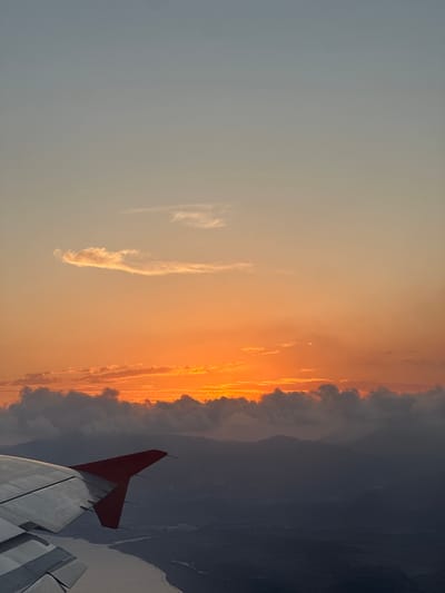 An aerial view from an aircraft captures a vivid sunset over a mountainous terrain near Dalaman, Turkey. The sky transitions from a pale blue-gray at the top, through warm orange mid-tones, to an intense, fiery orange glow at the horizon, where the sun is setting. A prominent, elongated cloud formation is illuminated by the sunset in the upper-mid section of the sky, with denser, dark clouds obscuring the lower portion of the sun's disc along the horizon. The left wing of the aircraft, rendered in silver-gray with visible panels and trailing edge devices, dominates the lower-left quadrant, featuring a distinct red wingtip. Below the wing, a light-toned body of water, likely a river, curves through a dark, shadowed landscape characterized by undulating hills and distant mountains, which appear silhouetted against the bright horizon. No people are visible.