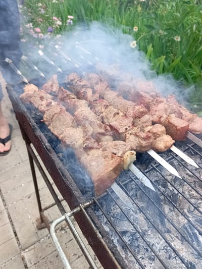 The image shows a close-up of several skewers of meat cooking on a grill. The meat is cut into cubes and arranged on metal skewers, with smoke rising from the grill. The grill itself is a rustic, metal construction, showing signs of rust and use. Beneath the grill grates, hot coals are visible, contributing to the smoke. In the background, out of focus, there is greenery with some small flowers, suggesting an outdoor setting. To the left of the grill, a person's leg and foot are visible, wearing a flip-flop, indicating a casual outdoor activity. The overall scene suggests a barbecue or outdoor cooking event. The lighting suggests it is daytime, and the presence of smoke implies the grilling is in progress. The context provided places the scene in Vyazovka, Russia. No text is visible in the image.