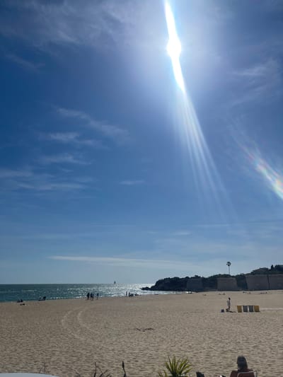 This photo captures a sunlit day at a beach in El Puerto de Santa María, Spain. A wide sandy beach stretches along the right, meeting the ocean on the left. The clear, blue sky is marked by wispy clouds and a bright sun. People are visible along the shore and in the water. Some beach chairs are placed near a series of small structures lining the edge of the beach. The sun's rays are prominent in the sky, creating a vibrant scene.