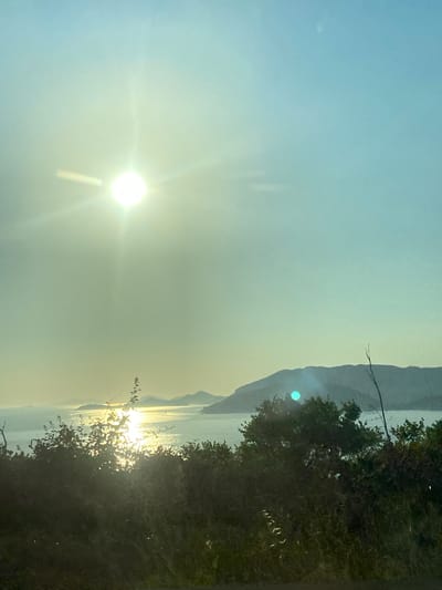 A wide shot captures a coastal view from a slightly elevated vantage point, overlooking the Adriatic Sea with islands in the distance. The sun, a bright orb in the upper left quadrant, casts a strong glare, creating lens flare and a hazy effect across the sky and water. The sky transitions from a pale blue at the top to a yellowish hue near the horizon. In the foreground, silhouetted bushes and grass create a dark, textured border along the bottom of the frame. The middle ground features the shimmering surface of the sea reflecting the sunlight, with several mountainous islands dotting the horizon. The overall atmosphere is serene and bathed in the warm light of the sun, likely during sunrise or sunset. The location is identified as Plat, Croatia.