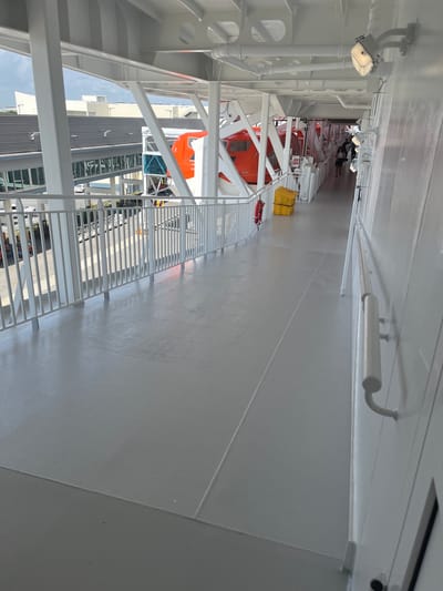This is a photograph taken from the deck of a ship, looking outwards. The perspective is rotated 90 degrees clockwise, as if the ship is tilted.
The subjects are primarily the ship's deck and its safety equipment. There are several orange lifeboats visible, suspended from davits. A yellow life raft is also present, along with a red life buoy. A white metal railing runs along the edge of the deck, casting shadows on the light grey surface. Several white pipes and lighting fixtures are mounted on the underside of the deck above, or on the ship's structure. In the distance, a building with a dark, ribbed facade and large windows can be seen. A few people are visible in the distance, near the lifeboats, though their details are not clear.
The scene appears to be outdoors on a bright day, suggested by the strong shadows and the clear sky visible in the distance between the building and the ship's structure. The lighting suggests it's daytime, possibly morning or afternoon. There is no visible text in the image. The overall impression is one of a functional, clean, and well-maintained part of a ship, equipped for safety and passenger transport.
