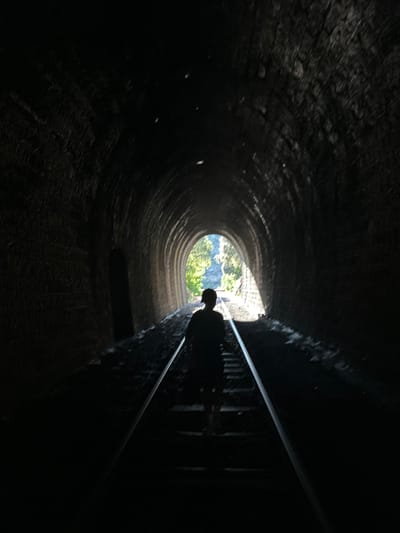 A person walks away from the camera down a railway track inside a dark, arched tunnel. Sunlight streams in from the tunnel's far end, illuminating the silhouette of the person and the tunnel's rough, stone walls. The person appears to be wearing a cap. The tunnel is approximately 150 meters long and is part of the Chemin de Fer du Train Touristique de l'Andouillette, located in Générargues, France.