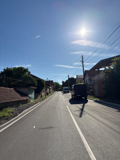 The image depicts a street scene in the city of Anton, Bulgaria, on a clear, sunny day. The perspective is from a vehicle traveling on an asphalt road with a white dividing line. On the left side of the road, there are houses with red tile roofs and some greenery. On the right side, there are more houses, one with a covered carport structure, and power lines running overhead. Two vehicles are visible on the road: a white van in the distance and a dark-colored van closer to the foreground. The dark van has a license plate that reads "EH 1116 MA". The sun is bright, casting strong shadows of power poles and the dark van onto the road. The sky is a clear blue with a few wispy clouds.