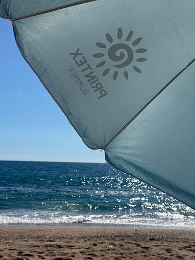The image displays a beach scene in Pineda de Mar, Spain, viewed from directly underneath a light blue beach umbrella. The umbrella's interior canopy, angled across the top of the frame and obscuring approximately two-thirds of the view, features a reversed dark grey logo reading "PRINTEX SUMMER" accompanied by a stylized sun icon. Below the umbrella, the deep blue Mediterranean Sea stretches towards the horizon, its surface shimmering with reflected sunlight and exhibiting small waves breaking into white foam at the shoreline. The foreground consists of a narrow strip of light brown sand marked with numerous footprints and indentations. A clear, light blue sky is visible above the sea on the upper left side of the frame. A small, indistinct white object is discernible on the distant sea horizon. No persons or explicit activities are depicted.