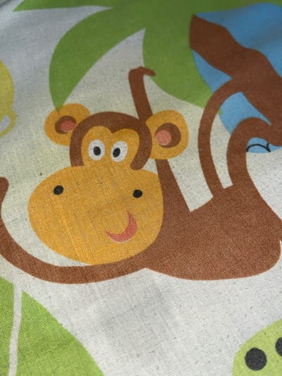 This is a close-up, high-angle, studio shot of a fabric print featuring cartoon monkeys and foliage. The fabric is a light, textured material, likely cotton or a blend. Dominating the foreground is the face of a monkey, rendered in bright yellow and orange, with black eyes and a red smile. Its brown arms and body are visible, with one arm reaching towards the viewer and the other extending upwards. The monkey is depicted swinging from a brown branch, set against a background of stylized green leaves and a blue shape, possibly representing the sky or another element of the jungle scene. The overall impression is of a playful and colorful design. There is no discernible location in Keudematangkuli, Indonesia, as this is an indoor studio shot of fabric.