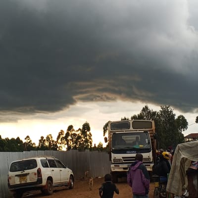 The image presents a scene in Keyian ward, Kenya, under a sky dominated by dark, ominous storm clouds with a sliver of bright sunset. A white Toyota van is parked on a dirt surface next to a corrugated metal fence. An Isuzu truck stands nearby, partially obscuring the view behind it. Two dogs are visible in the space between the car and the truck. In the foreground, several people are present: a child runs towards the truck, and others stand near a motorcycle, partially covered by a tarp. The atmosphere suggests an impending storm.