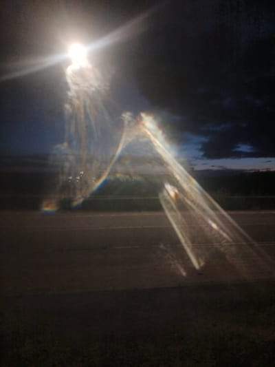 The image depicts a night scene with a bright light source, likely a street lamp, in the upper left quadrant. This light creates a significant lens flare and casts long, prismatic beams of light across the frame. The background consists of a dark sky with some clouds, and a horizontal road is visible in the lower half of the image. The overall impression is of a long exposure or a photograph taken with a lens that produces strong light artifacts. There are no discernable people or specific objects that can be identified with certainty due to the blurriness and light effects. The location in Воскресенское, Russia, is inferred but not visually confirmed by any landmarks within the image.