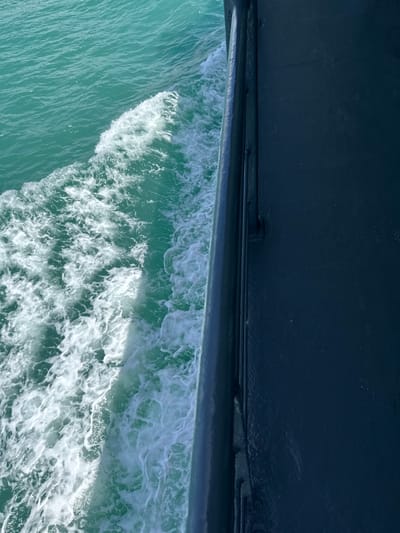 The image shows a dark blue hull of a boat against a bright turquoise sea with white foam created by the boat's movement. The perspective is from the boat, looking out at the water. There are no people visible in the image. The scene appears to be a snapshot of a boat traveling through the water, likely in a tropical or subtropical region given the color of the water. The location in Thong Nian, Thailand, is not discernible from the image itself; this information would be contextual.