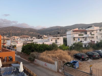 An elevated view overlooking a Mediterranean town towards a mountainous backdrop. White buildings with blue accents and air conditioning units are visible. Trees and dry brush add greenery. A restaurant with white umbrellas and outdoor seating is at the upper right. Parked cars and trash bins line the street to the left. The sky is a light blue, suggesting early morning or late afternoon.