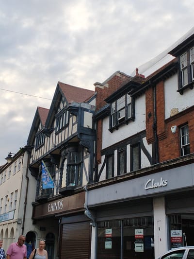 The image depicts a street scene in Wychavon, United Kingdom, featuring traditional architecture alongside modern storefronts. The buildings display a mix of styles, including Tudor-style timber framing and brickwork. On the left, a row of buildings with arched windows is visible. Further to the right, a building with prominent black timber framing and white plaster walls stands out. Above this, a blue flag with a floral pattern is displayed. The F. HINDS shop sign is visible on a dark brown background. Adjacent to it, the Clarks shoe store has a modern gray facade with its logo. Several signs are posted in the windows of the Clarks store, indicating relocation. Two individuals are in the foreground, walking from left to right. One, a man with a pink polo shirt and sunglasses, is on the left. To his right is a woman wearing a striped tank top. The sky is overcast.