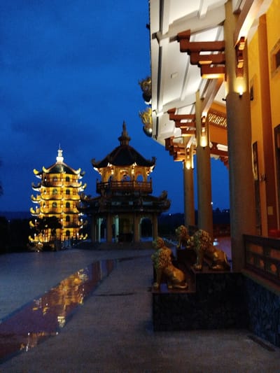 The image depicts a pagoda complex at night in Phú An, Vietnam. Two illuminated pagoda structures are visible in the background, one a multi-tiered tower. In the foreground, golden lion statues stand on a low stone platform near a building with prominent columns and overhanging eaves. The night sky is a deep blue. Reflected light is visible on the ground.