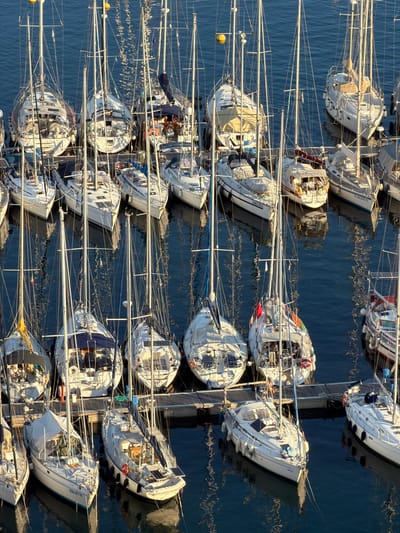 This is an aerial, high-angle, full shot of a marina in Santa Maria Navarrese, Italy, filled with numerous sailboats docked in calm, blue water. The sun is casting long shadows, indicating it is either early morning or late afternoon. The boats are white with various colored accents and sails, some furled and some loosely draped. Several boats have protective covers over their booms and cockpits. Some boats are docked side-by-side, while others are moored individually. A wooden dock runs along the bottom of the frame, with a few people visible on the boats, appearing relaxed and engaged in conversation or tending to the boats. One boat in the foreground has a license plate that reads "15 GE 0167 D". The water reflects the sky and the boats, creating a shimmering effect. The overall scene conveys a sense of tranquility and leisure associated with sailing.