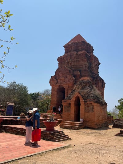 The scene depicts the Po Sah Inu Cham Towers in Phan Thiet, Vietnam, under a clear, bright blue sky. A large, reddish-brown brick Cham tower dominates the center-right, featuring a tiered structure, prominent ledges, and a pyramidal roof. A smaller, arched brick structure with a mound of dried vegetation on its roof is attached to the main tower's base, slightly to the right.
In the foreground, two adults stand on a reddish-brown tiled platform. The individual on the left wears a light-colored conical hat (nón lá), a dark sleeveless top, light trousers, and sandals, seemingly adjusting something on the other person. The second individual, on the right, wears a conical straw hat, a blue short-sleeved collared shirt, dark trousers, and flip-flops, holding a large red bag at their side.
Beyond these two, several other individuals are visible near the main tower's entrance. One person in a white shirt and dark pants is partially obscured while ascending brick steps into the tower. Another figure in a red shirt is discernible within the arched doorway.
A large, red terracotta pot containing a small plant with pink flowers sits on a low brick platform in the mid-ground. To the far left, a blue informational sign on a stand is partially visible, alongside sparse branches with light green leaves extending into the frame from the top-left. The ground transitions from the tiled platform to packed dirt and gravel surrounding the tower structures. Mature trees and shrubs with sparse foliage are visible in the background, suggesting a dry environment. The scene is well-lit by direct sunlight.