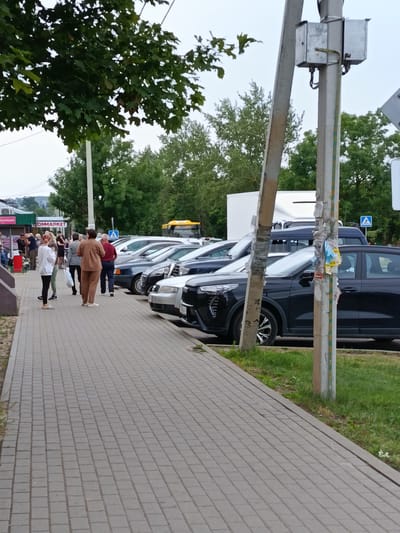 A group of people are gathered on a paved sidewalk in front of a row of parked cars. The cars are parked along the side of a street lined with trees. In the background, there are buildings and what appears to be a yellow bus. The overall scene suggests a public area, possibly a commercial or residential street, in the city of Smalyavitsky Sel'skiy Sovet, Belarus. The time of day appears to be overcast and possibly late afternoon or early evening.