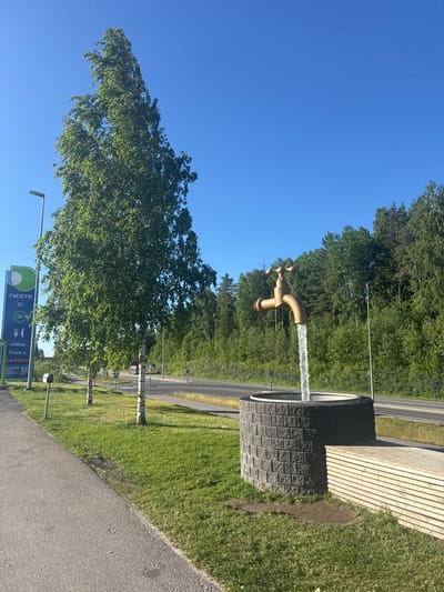 An outdoor scene in Heinola, Finland, features a large, golden faucet sculpture with water pouring from it into a circular stone basin. The sculpture is situated on a grassy area beside a paved walkway and a road. To the left of the scene is a tall, slender tree with green foliage, and further left, a tall sign for Neste, a Finnish energy company, is visible. The background is filled with a dense forest of green trees under a clear blue sky. The water from the faucet appears to be a continuous stream. A wooden bench is positioned to the right of the stone basin. The lighting suggests it is daytime, possibly late afternoon due to the warm glow on the sculpture and vegetation. No people are present in the image.