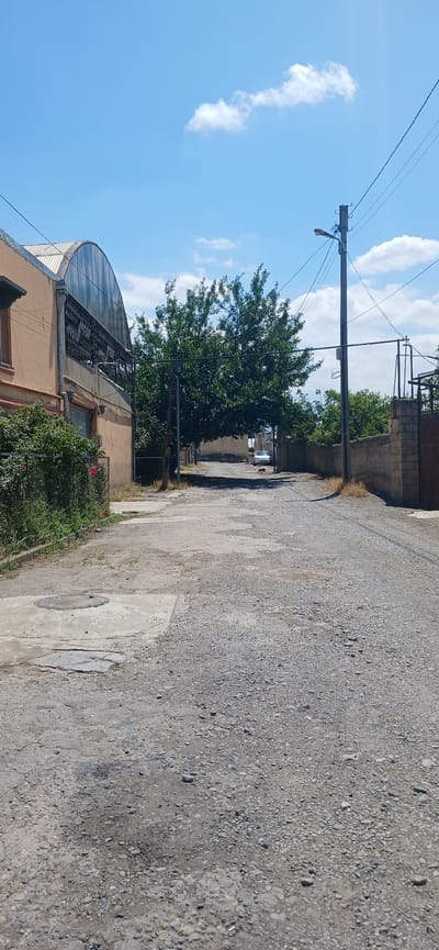 An upward-angle, medium-shot photograph taken on a bright, sunny day depicts a narrow, unpaved road lined with buildings and vegetation in Ganja City, Azerbaijan. The road surface is primarily composed of gravel and dirt, with visible patches of concrete and numerous potholes.
On the left side of the road, a light-colored building with a large arched, glass-paneled roof and ornate metal detailing is prominent. Adjacent to this building, a chain-link fence encloses a patch of green bushes and a single red rose. Further down the road on the left, a round concrete manhole cover is set into the ground.
Dominating the center of the frame is a large, mature tree with dense green foliage, casting a partial shadow onto the road. Beyond the tree, the road continues into the distance, narrowing slightly. A silver sedan is parked on the right side of the road, partially obscured by the tree.
On the right side of the road, a weathered concrete wall with some brickwork forms a boundary. A dark metal gate is visible at the far right edge of the frame. A utility pole with a street lamp and numerous overhead wires stands near the center of the road, close to the large tree. The sky above is a clear blue with scattered white, fluffy clouds. The scene appears to be a residential or semi-industrial area within Ganja City.