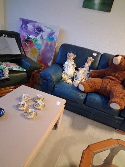 A blue leather sofa is topped with a large brown teddy bear and two porcelain dolls. Next to the sofa sits a light brown coffee table displaying four teacups and saucers. Beneath the sofa, a colorful abstract painting leans against a dark green chair with a floral cushion. A small mirror is positioned in front of the chair. A glass-topped wooden coffee table is in the upper right corner.