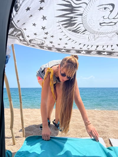 A woman with long blonde hair, sunglasses, and a yellow bandana tied around her neck is on a beach in Sant Pol de Mar, Spain. She is wearing a grey t-shirt and pink shorts and is bending down to adjust a turquoise beach towel on the sand. Behind her, the blue sea meets a clear sky. A patterned fabric, featuring a crescent moon and stars, is stretched between bamboo poles, creating a shaded area. The beach consists of small pebbles and sand.