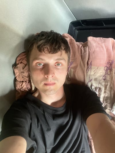 A first-person perspective video shows a man lying down, appearing to be in a train or bus. He is wearing a black t-shirt. The man has light skin, short brown hair, and green eyes. He is looking directly at the camera with a neutral expression. The background consists of a pink and purple patterned blanket and a black container. The setting appears to be indoors, with some ambient light suggesting it might be daytime. There is no visible text in the video. The man's head is resting on a folded pink cloth. The overall impression is casual and personal.