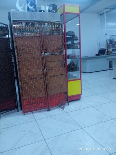 The image shows the interior of a commercial establishment, possibly a store, in Ak-Suu, Kyrgyzstan. In the foreground, a woven room divider with a red base is visible. To the left of the room divider, another dark woven screen is partially visible. Behind the room divider, shelves filled with various items, including what appear to be bottles and jars, are present. To the right, a tall, narrow display case with red and yellow framing stands, containing shelves with items that look like hats, bags, and possibly food products. In the background, a counter area with a cash register and a computer monitor is discernible. The floor is tiled. The timestamp "2026/6/30 08:03" is overlaid on the bottom right of the image. No people are visible in the scene.