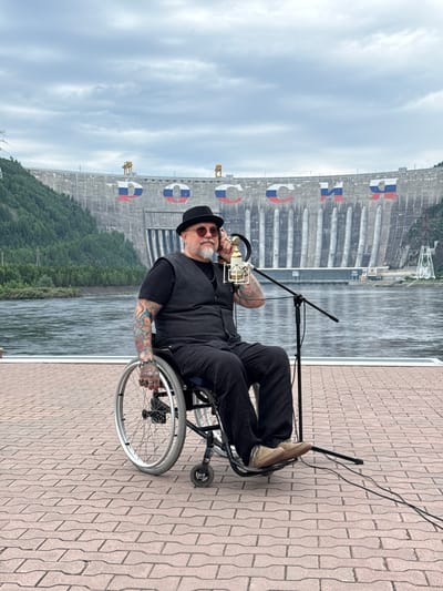 In Cheremushki, Russia, a man with tattoos sits in a wheelchair on a paved walkway. He wears a black hat, sunglasses, and black clothing. He is holding a microphone to his ear and speaking into another microphone positioned in front of him. Behind him is a large hydroelectric dam, the Sayano-Shushenskaya Dam. The dam is decorated with large letters painted in the colors of the Russian flag, spelling out "РОССИЯ" (RUSSIA). The sky is cloudy, and a river flows in front of the dam.