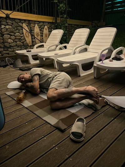 In Agronom, Russia, an adult male is asleep on a checkered blanket on a wooden deck. He is lying on his side, facing away from the camera, with his head resting on his arms. He is wearing a light-colored t-shirt and shorts. A plastic bottle of liquid and a white object, possibly a pillow or mat, are near his head. To his right, a flip-flop rests on the deck. In the background, there are several white lounge chairs, a stone wall decorated with three wooden surfboards, and some greenery. The overall scene suggests a relaxed, outdoor setting at dusk or nighttime.