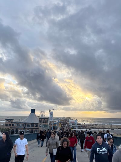 A crowd of people gathers on a pier with a Ferris wheel. Sunlight breaks through heavy clouds over the ocean. Many individuals stand near the edge, looking out at the water. A person wears an "Elon University" sweatshirt. Other figures stroll along the pier, enjoying the view. The "Santa Monica Pier" sign is visible.
