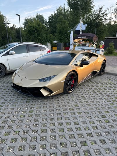 The image captures a golden Lamborghini Huracan parked on a permeable pavement in a semi-urban environment, likely in Barvikha, Russia. The car is positioned in the foreground, angled slightly towards the viewer, showcasing its sleek design and a large rear wing. Adjacent to the Lamborghini, a white hatchback is partially visible, suggesting a parking area. In the background, a food cart with the "MONTERRA" logo is set up, with a person visible behind the counter, serving customers. Trees and foliage surround the area, and a street lamp stands tall in the distance. The overall scene appears to be an outdoor gathering or a public space with commercial activity.