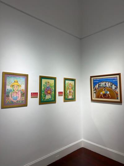The image shows a corner of a gallery wall in La Orotava, Spain, displaying four framed paintings. The walls are painted white, and the floor is dark wood.

From left to right:

The first painting features a woman wearing a wide-brimmed straw hat and a pink dress, holding a baby. She is surrounded by a vibrant floral background with trees. The frame is gold. Two small red placards are affixed to the wall to the right of this painting.

The second painting depicts a woman with a headscarf, holding an earthenware pot. She is standing in front of a background of green foliage and orange flowers. The frame is green and gold. A red placard is on the wall to the right of this painting.

The third painting shows a woman in a yellow headscarf and a white dress, holding an earthenware pot. She is positioned in front of green foliage with orange flowers. The frame is green and gold. This painting is smaller than the first two.

The fourth painting, in the far right corner, is larger and shows a scene of a group of people gathered in a semi-circle, watching two figures wrestling in the center. The figures in the wrestling ring are wearing blue and yellow singlets. The frame is wood with a darker wood inner border.