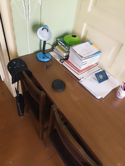 A desk in a room has a blue lamp, a stack of books, a passport, and a black hat hanging on a chair. The room appears to be in a residential setting in Aygezard, Armenia. The desk is made of wood and is positioned in front of a wall with a door. The floor is also made of wood and has a distressed finish. Two wooden chairs are positioned at the desk. A green decorative item is placed on top of some books. A small black circular object is also on the desk. A cup with pens and pencils is located on the right side of the desk. A black purse and hat are hanging from the back of a chair.