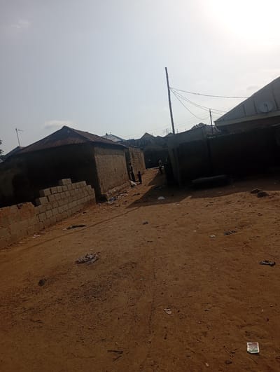 The image depicts a dusty, unpaved street in Gwagwa, Nigeria. Structures with corrugated metal roofs and mud or brick walls line the street, some appearing unfinished or in disrepair. The ground is covered in loose, reddish-brown soil and debris, including scattered plastic bags and other refuse. In the background, several figures, including at least two adults and one child, are visible near the buildings. Overhead power lines stretch across the scene, supported by wooden poles. The sky is bright and hazy, suggesting a sunny day.
