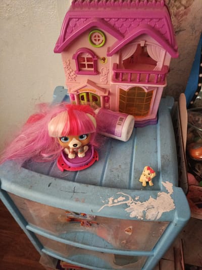 A pink dollhouse with multiple levels and balconies sits atop a blue plastic drawer unit. A doll with long pink hair, featuring a bipartite pink and white color scheme, stands on a pink base in front of the dollhouse. A small, light-colored toy dog with a pink bow is positioned on the top surface of the drawer unit, to the right of the dollhouse. The drawer unit is visibly dirty, with white paint splatters on its top surface and some smudges and scratches. A portion of a pink bottle or container is visible near the dollhouse. The background consists of a textured wall and some miscellaneous items to the right, including a dark blue and white patterned object and a clear plastic bag containing other objects. The scene appears to be indoors, likely a domestic setting. There is no indication of the city of Meget, Russia.