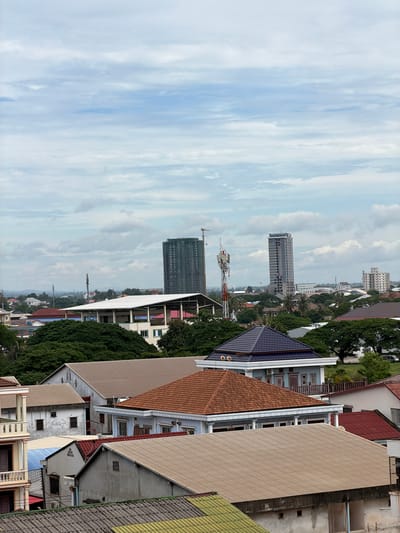 This is an aerial, eye-level view of a densely packed urban landscape in Vientiane Capital, Laos. The foreground is dominated by the roofs of several residential buildings, most with tan or grey corrugated metal roofing. One prominent building in the middle ground has a dark blue, multi-faceted roof with white trim. Behind these, a large, white, open-sided building with a slanted roof stretches horizontally.

Further back, several tall, modern buildings rise above the treeline, suggesting a mix of older and newer development. Two particularly tall skyscrapers are visible, one under construction with a crane. A prominent cell tower with red and white accents stands between two of the taller buildings. The sky is overcast with scattered clouds, obscuring a clear view of the sun. The scene depicts a built-up area with residential homes, commercial structures, and infrastructure typical of a developing city. There are no people or animals visible in the image.