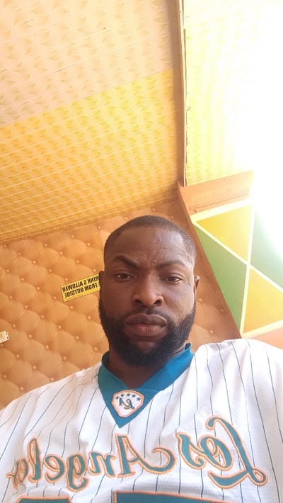 An adult male subject is depicted in a chest-up portrait within an indoor environment in Dandagoro, Nigeria. The individual, appearing to be of African descent, has a short, dark haircut and a trimmed beard. He wears a white pinstriped jersey with a teal collar and teal lettering, outlined in orange, spelling "Los Angeles" across the chest. A patch with "LA" and five stars is visible on the upper left chest area. The subject faces directly forward with a neutral expression. The background features a patterned ceiling composed of light and dark yellow panels with a wavy design. To the subject's left and behind him, a wall is covered in an orange-brown tufted or quilted material. A yellow rectangular sign with black, inverted text, stating "NO DRINKS ALLOWED FROM OUTSIDE," is visible on this wall. To the subject's right, a structural element or corner displays geometric patterns in green, yellow, and white, with brighter illumination present in this area.