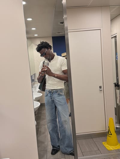 A young Black man is taking a mirror selfie in what appears to be a public restroom. He is wearing a white ribbed short-sleeve shirt, light wash wide-leg jeans, black loafers, and sunglasses. He has white earbuds in and is holding a smartphone in his right hand. The restroom has light grey tiled floors, white sinks, and white stall doors. There is a yellow wet floor sign visible in the reflection on the right. The man is standing in a location that is consistent with a public restroom, potentially within the city of Crawley, United Kingdom, given the presence of a wet floor sign which is common in such establishments.